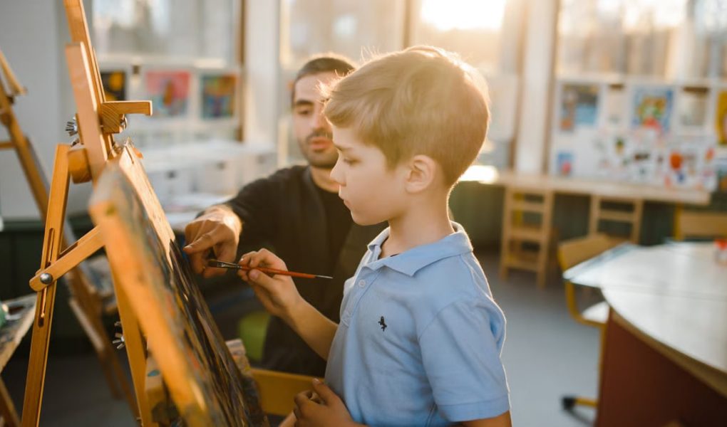 A young boy painting on an easel with a teacher assisting in a sunny art classroom.