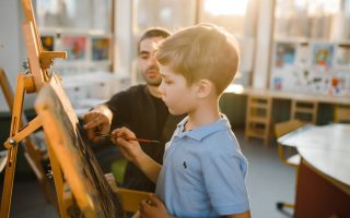 A young boy painting on an easel with a teacher assisting in a sunny art classroom.