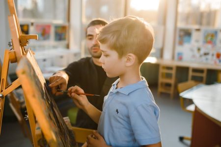 A young boy painting on an easel with a teacher assisting in a sunny art classroom.